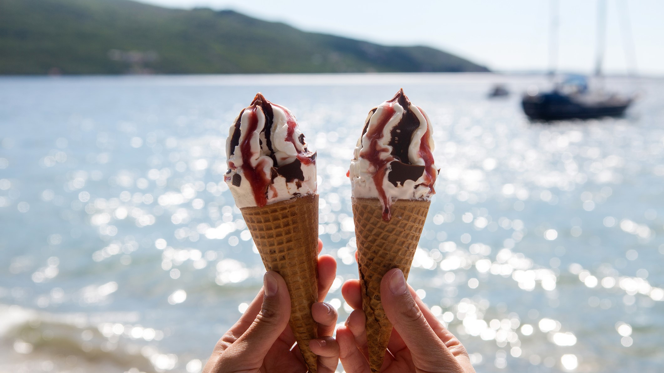 hands holding ice cream cones near a body of water